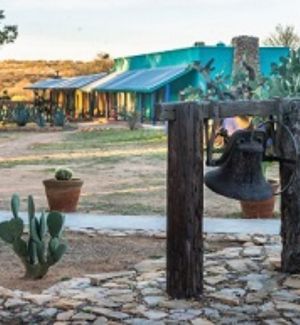 Photo of a scene with prickly pear cactus, pathways to a turquoise villa in the background and a large bell 